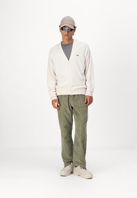 Lys beige cardigan med v-hals og knaplukning, parret med olivengrønne kordfløjl bukser og lyse sko.