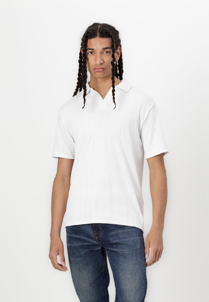 Vit kortärmad polo med ett texturerat mönster och V-halsringning. Matcher med mörkblå jeans, stående framför en enfärgad bakgrund.