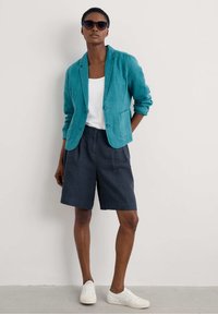 Teal linen blazer over a white tank top, paired with dark shorts and white sneakers. Features buttons, one pocket, and round black sunglasses.