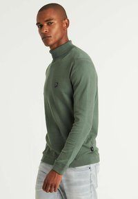 Grüner Pullover mit hochstehendem Kragen, strukturiertem Stoff und einem kleinen schwarzen Emblem auf der Brust. Enganliegendes Design mit gerippten Bündchen und Saum.
