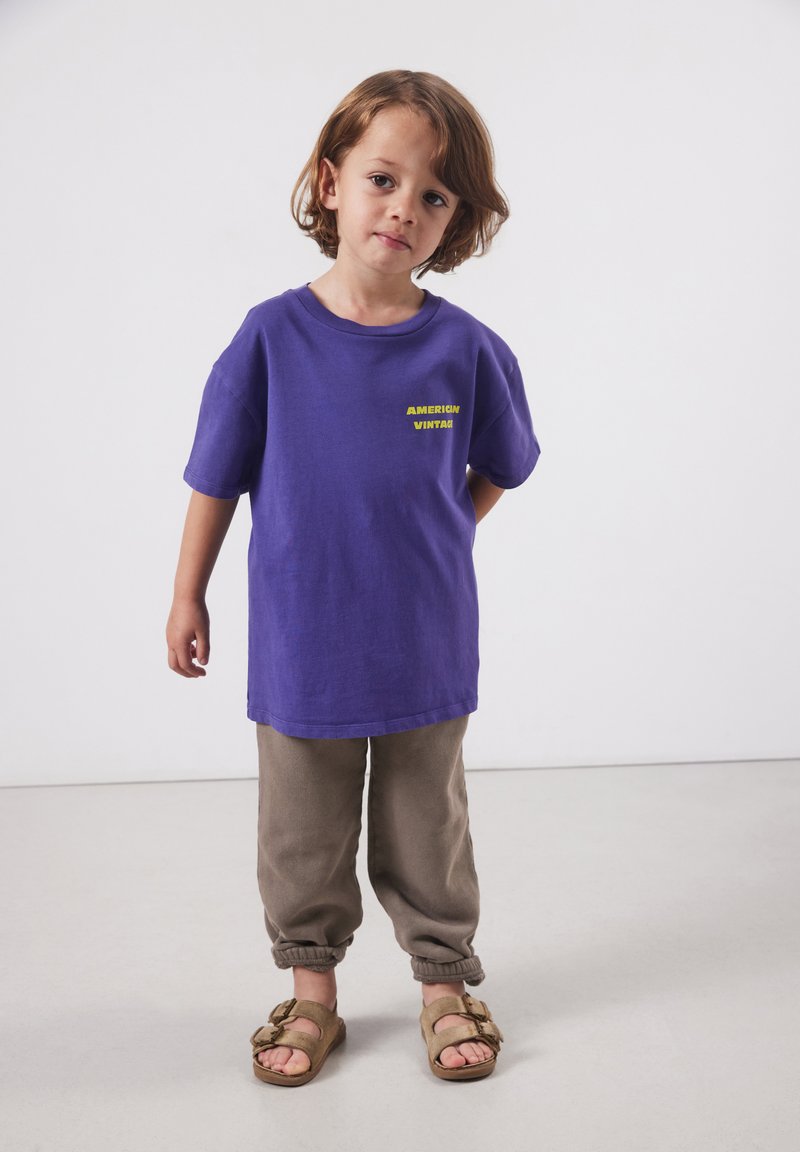 Jong kind staand, draagt een paarse oversized T-shirt met gele tekst "American Vintage", beige joggingbroek en beige sandalen.