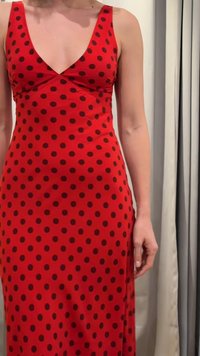 Γυναίκα που φοράει ένα αμάνικο κόκκινο φόρεμα με μαύρες πουά και βαθύ V-λαιμό, στέκεται δίπλα σε ανοιχτόχρωμες κουρτίνες.