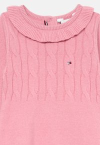 Svetlo roza pletena obleka z vzorcem kabelskega pletenja. Ima rebrast ruffle ovratnik in majhen logotip na sprednji strani. Mehek material.