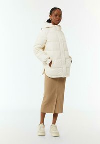 Crème pufferjack met capuchon, voorkeurzakken en drukknoopsluiting, gecombineerd met een geribbelde beige midi-jurk met een centrale knoopsluiting.