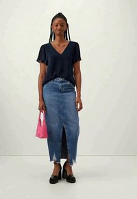 Ung kvinne står iført en svart V-hals blouse, blå denimskjørt med lysken foran, svarte platåhæler, og holder en liten rosa håndveske.