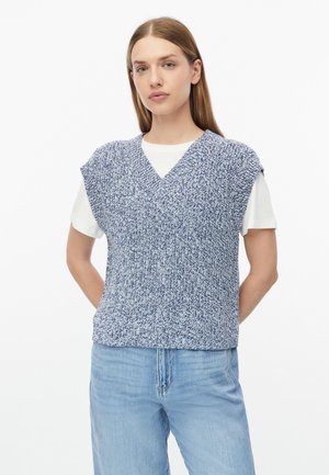 Giovane donna con capelli lisci che indossa un maglione smanicato lavorato a maglia blu e bianco sopra una T-shirt bianca e jeans azzurri chiari.