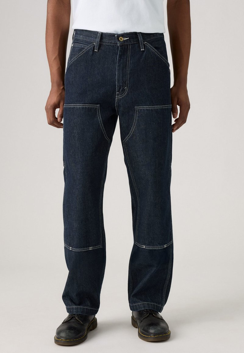 Muškarac u tamnoplavim trapericama s kontrastnim šavovima i crnim kožnim cipelama, stoji protiv jednostavne pozadine.