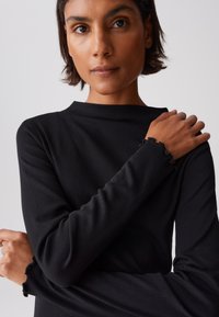 Schwarzes Langarmshirt aus einem weichen Stoff mit gerippter Textur. Hat einen hohen Kragen und Rüschen an den Manschetten beider Ärmel.