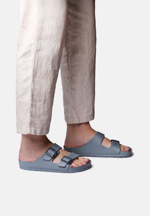 Person iført beige bukser og grå slip-on sandaler med to justerbare spænder, stående på en ensfarvet hvid baggrund.