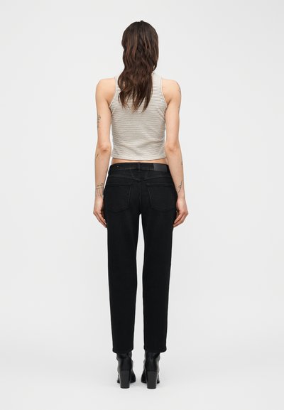 Femme aux longs cheveux ondulés portant un haut rayé sans manches, un jean noir à jambes droites et des bottines à talons noires, vue de dos.