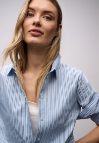 Jeune femme aux longs cheveux blonds portant une chemise à rayures bleu clair sur un haut blanc, regardant la caméra avec assurance sur un fond uni.