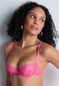 Femme aux cheveux bouclés portant un soutien-gorge en dentelle rose vif et des boucles d'oreilles en métal doré, regardant vers sa droite devant un fond clair.