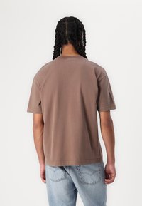 Brown cotton T-shirt with short sleeves, crew neckline, relaxed fit, and a smooth texture. Paired with light blue denim jeans.