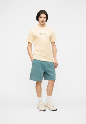 Νέος άνδρας στέκεται με τα χέρια στις τσέπες, φορώντας μπεζ μπλουζάκι με σχέδιο, τιρκουάζ σορτς, άσπρες κάλτσες και μπεζ αθλητικά παπούτσια σε απλό φόντο.