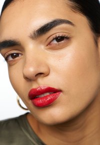Close-up of a face featuring smooth, medium-toned skin, defined eyebrows, and glossy red lips. Hair slicked back, with large hoop earrings.