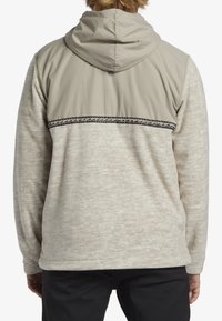 Lekki beżowy hoodie z teksturowanym kremowym korpusem i gładkim brązowym yoke, ozdobiony czarnym wzorowanym akcentem na ramionach.