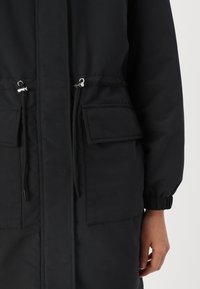 Cappotto imbottito nero con chiusura frontale a zip, vita regolabile con coulisse, polsini elastici e due tasche a toppa sul davanti. Tessuto con texture liscia.