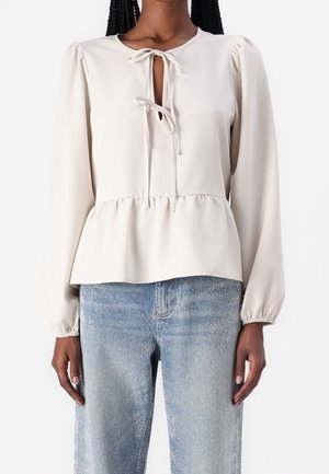 Woman wearing a cream long-sleeve blouse with front ties and gathered hem, paired with light blue patterned denim jeans.