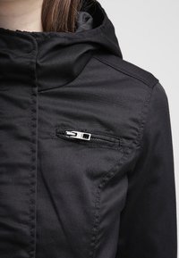 Czarna kurtka z kapturem wykonana z teksturowanego materiału, z wyraźnymi przeszyciami oraz kieszenią na piersi zapinaną na suwak z srebrnym okuciem.