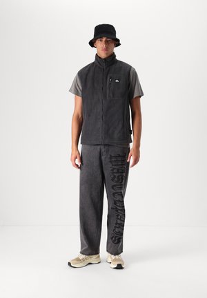 Gray fleece vest with a zip pocket, paired with gray textured pants featuring black graphic text. Black bucket hat and cream sneakers complete the outfit.