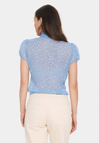 Himmelbluse mit floralem Muster, kurzen Puffärmeln, gerafftem Kragen und transparenter Textur, kombiniert mit hellbeigen Hosen.