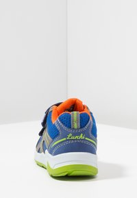 Zapatilla para niño en azul, gris y naranja con detalles en verde, tiras de Velcro y suela blanca, vista desde atrás sobre una superficie blanca.