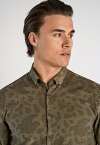 Gebutton-up overhemd in olijfgroen, gemaakt van textuurstof, met een ingewikkeld paisleypatroon en zwarte knopen aan de voorkant.