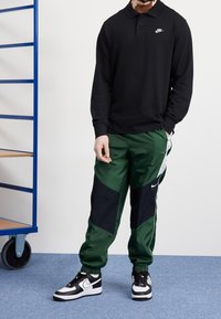 Osoba ubrana w czarną, długą koszulę z logo Nike, zielono-czarne spodnie dresowe Nike i czarno-białe sneakersy Nike stoi na niebieskim dywanie.