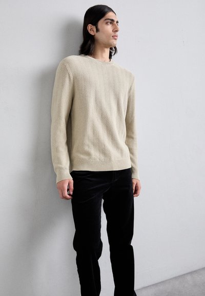 Pull en laine beige avec un motif en chevrons texturé, encolure ronde et poignets côtelés, associé à un pantalon en velours côtelé noir.