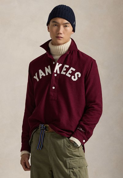 Μαραoon μακρυμάνικο πουκάμισο με γράμματα "YANKEES", κουμπωτή κολάρο, στρωμένο με κρέμα λαιμόκοψη. Φορεμένο με ελαιόχρωμες cargo παντελόνες και ναυτικό σκουφάκι.