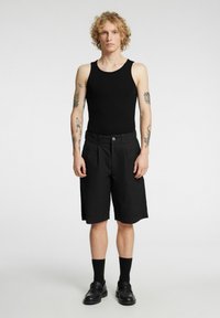 Czarny top bez rękawów i czarne plisowane szorty. Buty to czarne mokasyny. Modelka ma kręcone włosy i widoczne tatuaże. Jednolite tło.