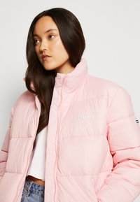 Rosa puffer-jakke med høy krage, med en hvit brodert logo på brystet. Stoffet har en glatt tekstur og kviltet mønster.