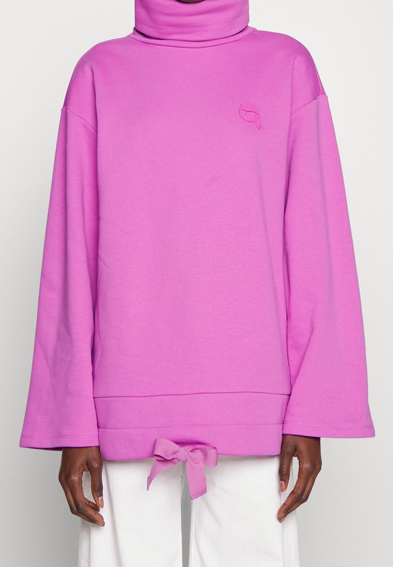Sudadera rosa de gran tamaño con cuello alto, mangas anchas y un detalle atado en la parte inferior. Presenta un sutil logotipo en relieve en el pecho.