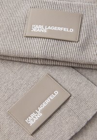 Pilkas megztas audinys su tekstūriniu raštu, turintis stačiakampį šviesiai rudos odos atlapą su užrašu "KARL LAGERFELD JEANS" baltos spalvos.