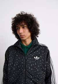 Black zip-up jacket with a leopard print pattern and white Adidas logo, paired with a green polo shirt, featuring textured fabric and stripes.