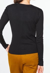 Person som bär en svart långärmad tröja och senapsgula byxor, står med en hand i fickan, vänd bort från kameran.