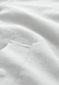 Tejido gris claro con una textura suave, que presenta sutiles detalles de costura y un pequeño bolsillo. El material parece suave y transpirable.