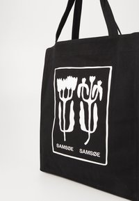 Zwarte stoffen draagtas met een witte bloemenafbeelding en de tekst "SAM SøE" onderaan. Stevige handvatten voor gemakkelijk dragen.