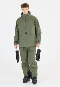 Olivgrön skidjacka med halvklar dragkedja och svarta detaljer, tillsammans med matchande byxor. Svarta handskar hålls i ena handen. Vattentätt material.