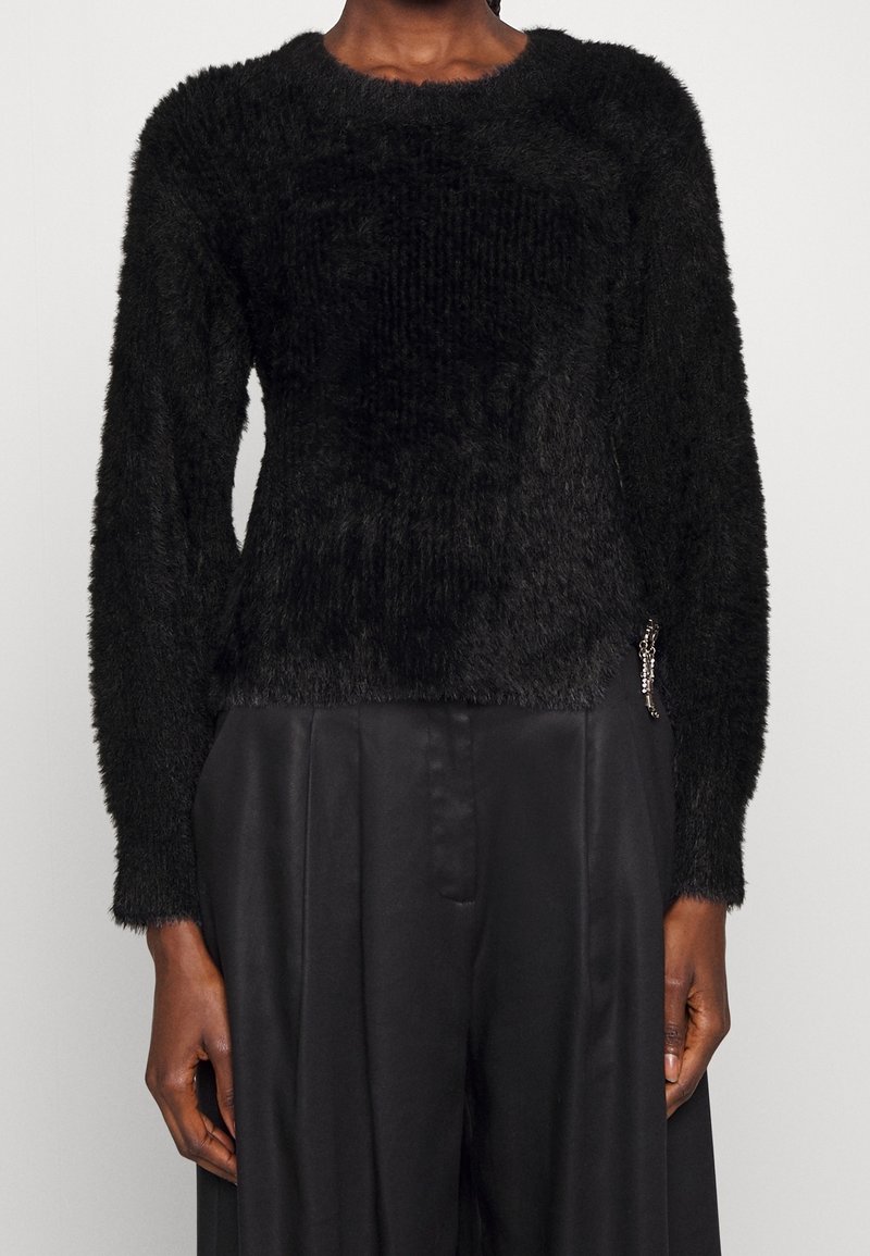 Kvinna i en svart fluffig långärmad tröja, som har lösa svarta satinpants, stående mot en neutral ljus bakgrund.