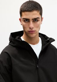 Schwarze Zip-up-Jacke mit Kapuze, die eine glatte Textur und ein minimalistisches Design aufweist. Über einem weißen T-Shirt getragen. Nahaufnahme-Porträt.