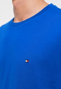 Karaļa zila kokvilnas t-kreklu ar apaļu kakla izgriezumu. To rotā neliels logotipa uzlīme sarkanā, baltā un melnā krāsā uz kreisā krūšu laukuma. Mīksta tekstūra.