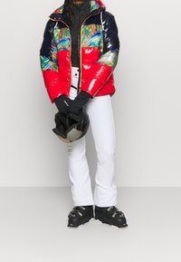 Veste rouge rembourrée avec un design coloré, accents noirs ; gants noirs ; casque noir ; pantalon de ski blanc ; texture et coupe élégantes adaptées au ski.
