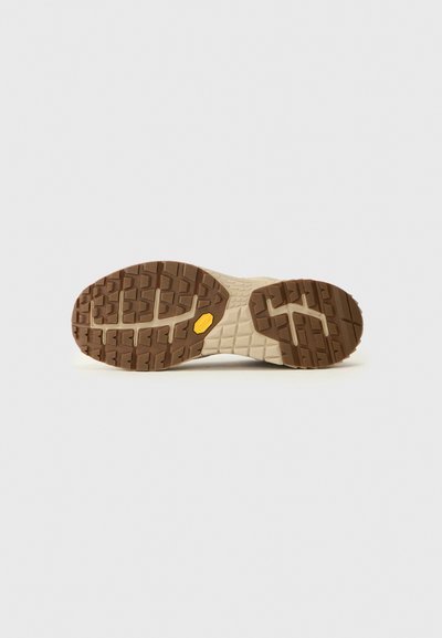 La semelle extérieure des chaussures présente un motif de bande en caoutchouc brun avec des crampons profonds et une midsole beige texturée, mise en valeur par un logo de marque jaune proéminent.