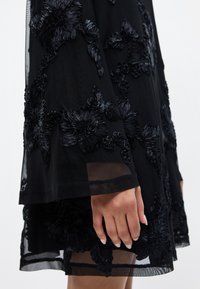 Melnas kleitas ar sarežģītu ziedu izšuvumu, caurspīdīgu audumu, zvana piedurknēm un teksturētu apdari, izceļot izsmalcinātu amatniecību.