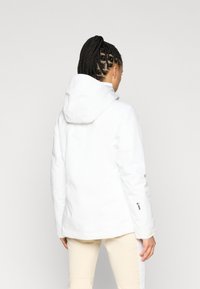 Mulher com cabelo trançado, vestindo uma jaqueta branca com capuz e calças bege claro, de costas contra um fundo claro e simples.