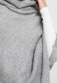 Personne portant un châle gris scintillant en tricot sur un haut blanc à manches longues, tenant le châle près du cou avec une main.