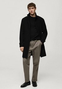 Svart kappa med hög krage, bärs över en svart tröja och beige byxor. Modellen står i svarta skor mot en neutral bakgrund.