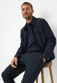 Marineblauwe lichtgewicht jas met twee voorzakken, gecombineerd met een bijpassend marineblauw t-shirt en broek, weergegeven op een houten kruk.