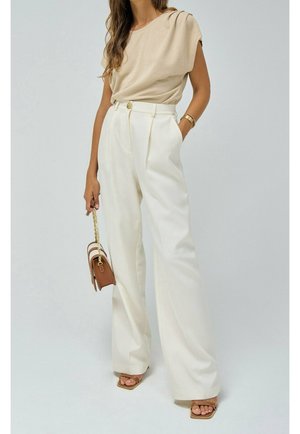 Frau trägt ein beiges Kurzarm-Top und hochtaillierte weiße weite Hosen, hält eine kleine braune Handtasche mit goldener Kette.